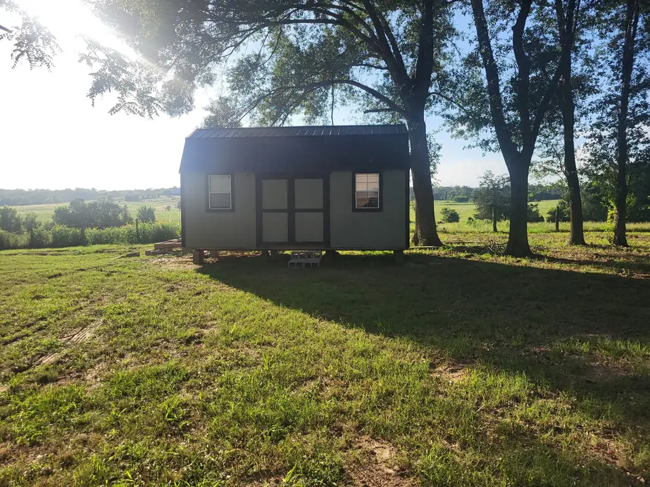 A small cabin under tall shade trees, mown grass in front and open pasture rolling away behind