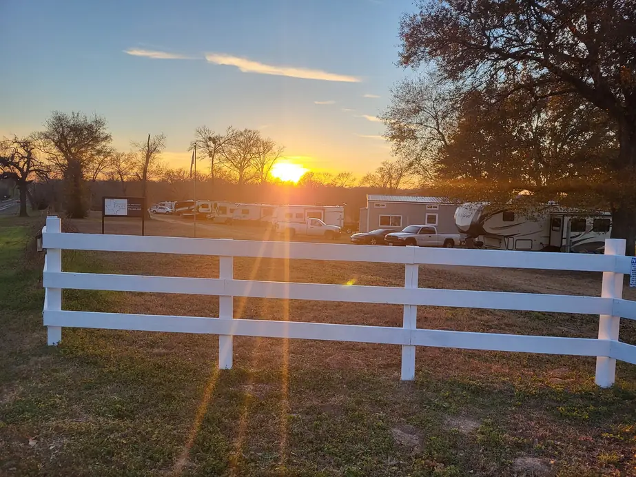 A white three-rail ranch fence at the front of the park at sunset, RVs and trucks parked under big oaks behind it