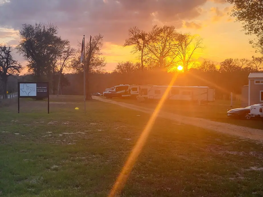 Golden-hour sun over a row of fifth wheels and travel trailers on the gravel drive, the park's Texas-map sign and flagpole at left
